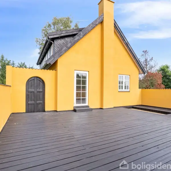 Gul bygning med sort tag, placeret ved stor træterrasse. Omgivet af træer under en blå himmel. Mørk dør og vinduer med hvide rammer ses i midten.