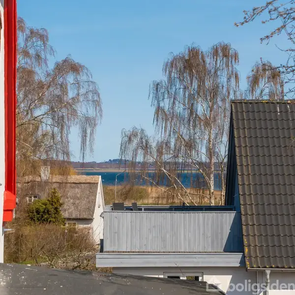 Bygninger med røde og grå tage står stillet i en forstad. Bag dem ses høje træer og en glimt af havet. Solen skinner, og himlen er klar blå.