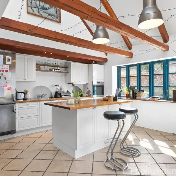 Køkken med hvid køkkenø, to barstole og rustfrit stål køleskab, omgivet af hvide skabe. Træloftsbjælker og lysekranser hænger over. Store vinduer slipper lys ind. Tiled gulv og moderne indretning.