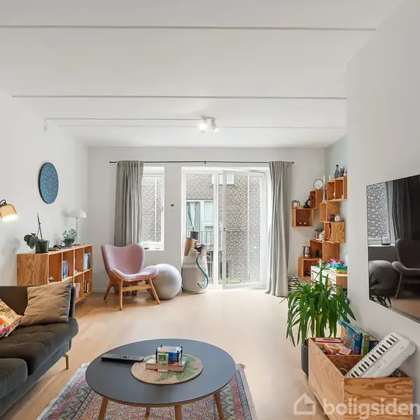 Stue med en sort sofa fyldt med farverige puder. Et rundt sofabord står foran sofaen. Store vinduer med gardiner i baggrunden, som giver naturligt lys.