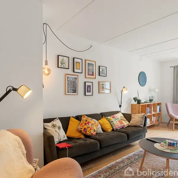 I en stue står en mørk sofa med farverige puder, omkranset af vægkunst og lamper. Et rundt sofabord ligger foran, omgivet af komfortable stole og reoler ved vinduer.