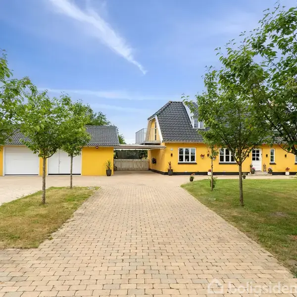 Gul villa med mørkt tag og to etager står for enden af en stenbelagt indkørsel. Træer flankerer stien, og huset ligger i et landligt område under en blå himmel.