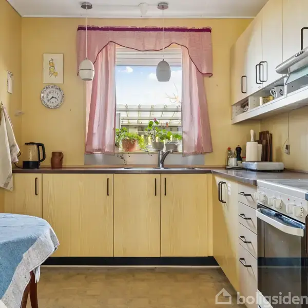 Et køkken med lyse træskabe og grå bordplader. En ovn og komfur står til højre, mens en vask er under et vindue med lyserøde gardiner og planter. Bord med blå dug til venstre.