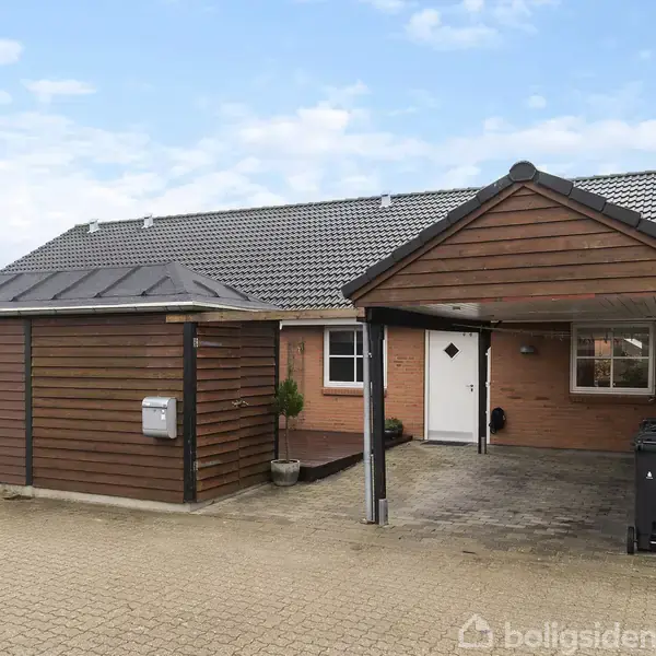 Rækkehus med skråt tag og carport, omgivet af flisebelagt gårdhave. Der er to postkasser på væggen og affaldscontainere ved indkørslen. Himlen er delvist skyet.