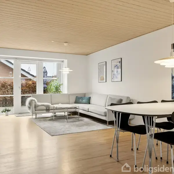 En lys stue møbleret med en grå hjørnesofa og et seks-personers spisebord. Vægge dekoreret med kunst, store vinduer med udsigt til en have. Træloft og parketgulv.