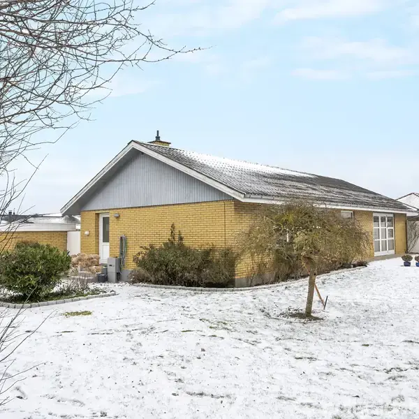 Et gult murstenshus er omkranset af en snedækket have. Træer og buske er delvist dækket af sne, mens et lyst, overskyet himmelstrækning ses i baggrunden.