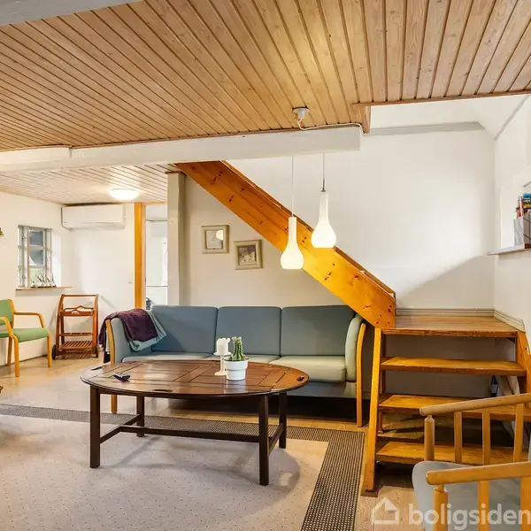 En stue med en blå sofa står foran en trappe. Et sofabord med planter er i midten, omgivet af stole. Rummet har lyse vægge og et træloft.