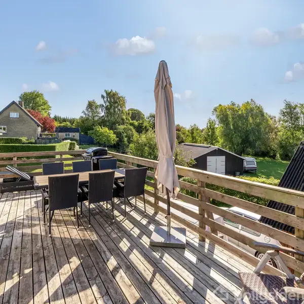 En udendørs terrasse med træmøbler og en parasol står under en klar blå himmel. Omkring terrassen er der grønne haver og huse i baggrunden.