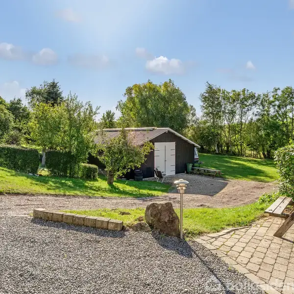 En træbygning med åbne hvide døre står blandt grønne træer og buske. Foran er en grusvej og en flisebelagt gårdhave med et picnicbord på en solrig dag.