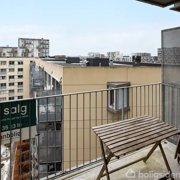 En altan med et bord og to stole står over en byudsigt. Et skilt med teksten "Til salg, danbolig, 35 35 23 10" hænger på rækværket.