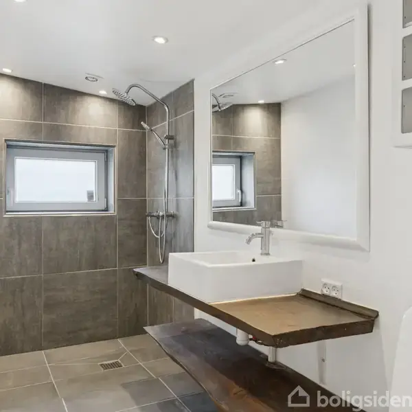 Et moderne badeværelse har en bruser med glasdør til venstre, en hvid håndvask monteret på en mørk bordplade, et stort spejl over vasken, grå fliser og en lille firkantet vindue.