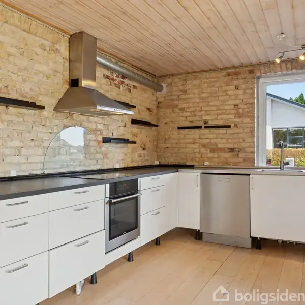 Hvidt køkken med sorte bordplader. Der er en ovn og emhætte. Rummet har en murstensvæg og et vindue med udsigt til en have.