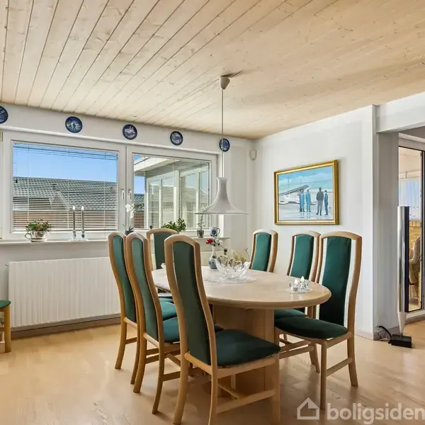 Rundt spisebord med grønne stole står i spisestue med lyst trægulv og loft. Vinduer viser udsigt til solpaneler. Billede hænger på væggen, dekorative tallerkener over vinduet.