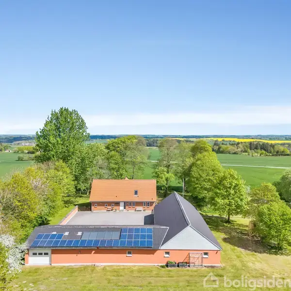 To huse med solpaneler på taget står i et åbent landskab omgivet af grønne marker og træer under en klar blå himmel.