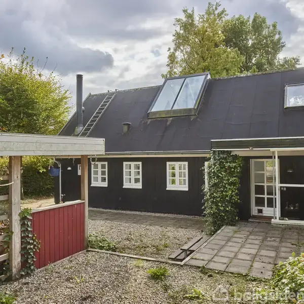 Sort hus med tagvinduer ligger i en gårdhave omgivet af træer og planter. Der er en stige på taget, flisebelagt terrasse og grusbelagt gårdsplads.