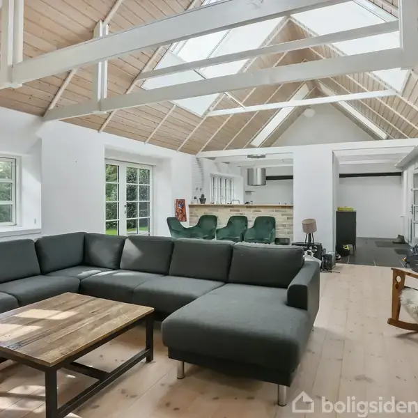 Stor, mørkegrå hjørnesofa står i en lys stue med skråloft og ovenlysvinduer. Foran sofaen er et træbord, og der er en gyngestol ved siden af. Baggrunden viser et åbent køkken.