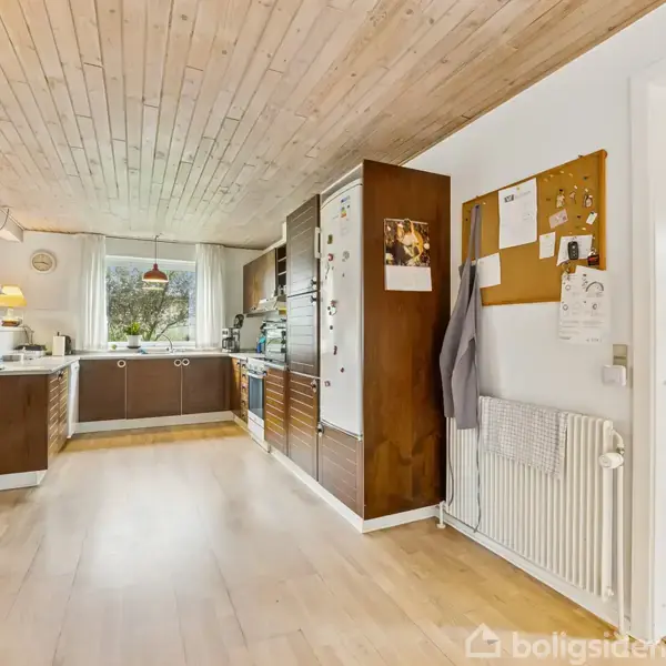 Køkken med brunlige træskabe har køleskab ved væg med opslagstavle. Ophængte lys over bordet. Trægulv strækker sig til åbent tilstødende rum. Vandkedel på bord, vindue med udsigt til have.