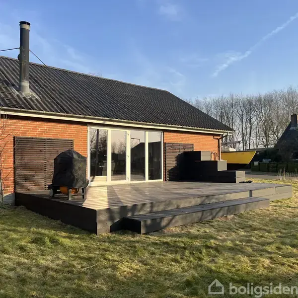 En træterrasse med grill og siddepladser foran et rødt murstenshus. Store glasdøre åbner mod terrassen. Græsplæne og træer i baggrunden.