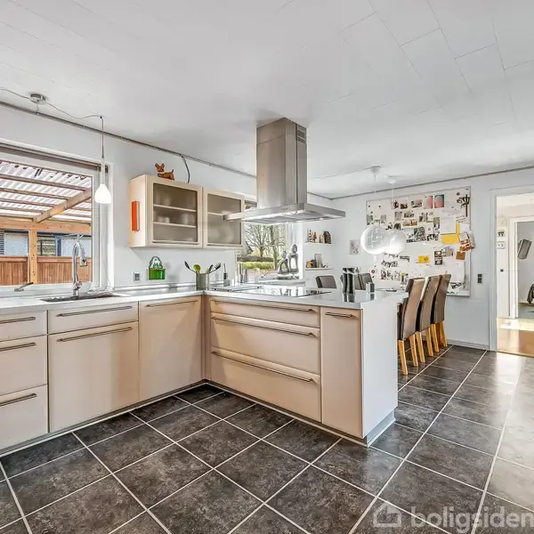 Køkken med lyse skabe og en kogeø, omgivet af hvide vægge og et vindue mod en terrasse. Til højre er en spiseplads med seks stole. Mørkt flisegulv.