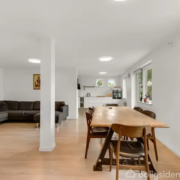 En spisestue med træbord og seks stole står i en lys, moderne stue med trægulv. I baggrunden ses et åbent køkken og en hjørnesofa.