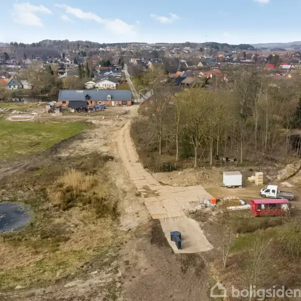 Byggeplads med et betonvej nær et skovområde, omkranset af byggeudstyr som en lastbil og containere. I baggrunden ses en by med huse og træer.