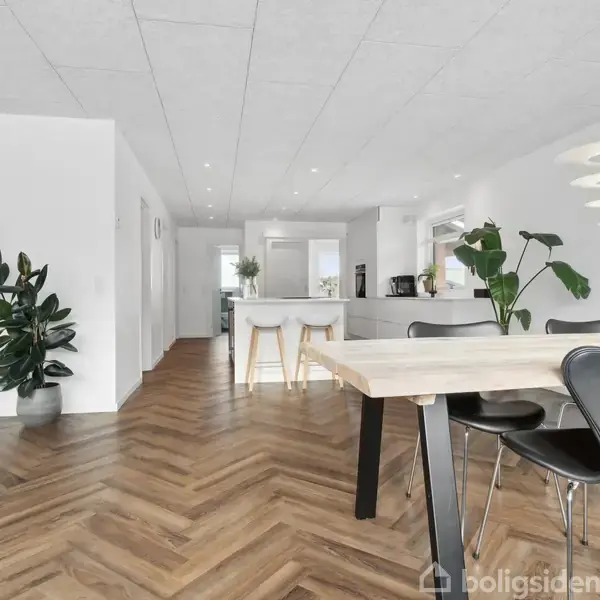 Spisebord med sorte stole står i et moderne, lyst køkken-alrum. Trægulv i sildebensmønster, hvide vægge, væghængt fladskærm og planter skaber en indbydende atmosfære.