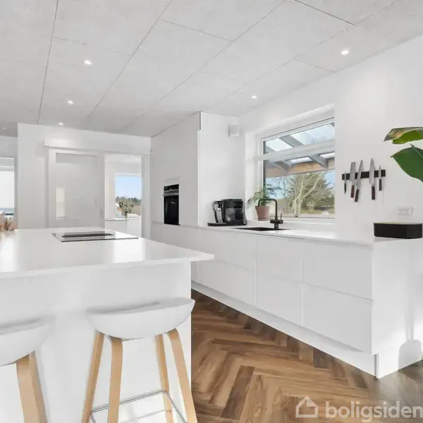 Et moderne køkken med hvidt interiør og trægulv. En plante står i hjørnet, og en ø med to barstole er centralt placeret. Blomster og køkkenudstyr pryder rummet.