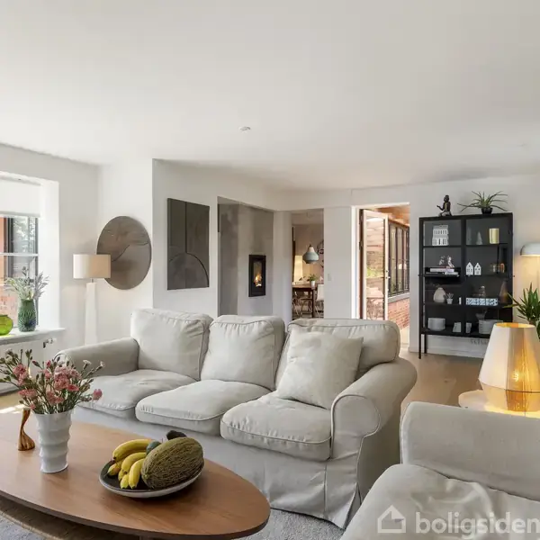 En stue med hvide sofaer står stille, omgivet af en moderne indretning med grønne planter og kunst. Et træbord bærer frugter og blomster. Store vinduer slipper lys ind i rummet.
