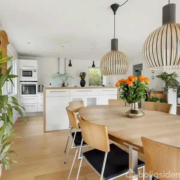 Et rundt spisebord med en vase fyldt med orange blomster står centralt. Køkkenet i baggrunden har hvide skabe og moderne apparater, mens store lamper hænger over bordet. Planter pynter rummet.