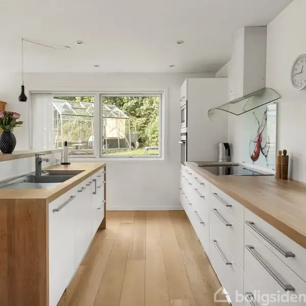 Køkken med hvide skabe og træbordplader; vask og kogeplade er indbygget. Et stort vindue giver udsigt til en have med et drivhus. Vaske og blomster ses på bordet.