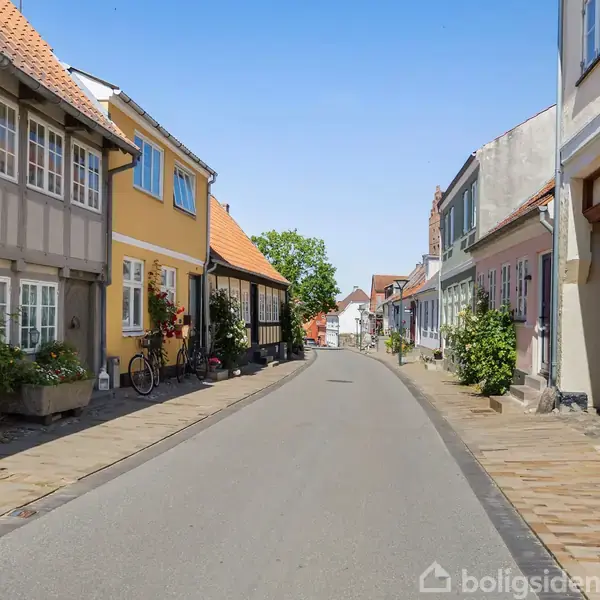 Farverige, traditionelle huse står langs en snoet vej i en rolig by. Cykler og grønne planter ses på fortovene, mens blå himmel indikerer klart vejr.