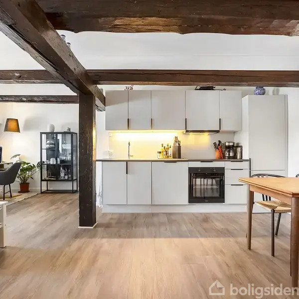 Et moderne køkken med hvide skabe og indbygget ovn. En spiseplads med træbord og stole står ved siden af. Rummet har synlige bjælker og trælaminatgulv, og der er indrettet en stue med lamper og planter.