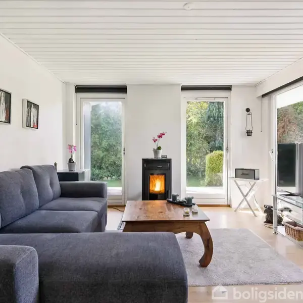Grå sofa står i stue med brændeovn under to store vinduer. Der er et sofabord, billeder på væggen, og en TV på en stander.