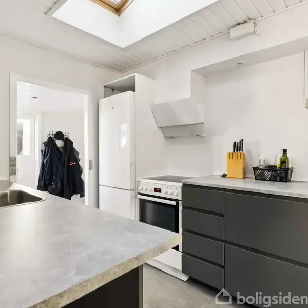 Køkken med mørkegrå skabe, hvide vægge, og lys bordplade. Komfur ved siden af køleskab, knivblok og olier på bordet. Skylight og åben dør mod gang med knagerække.