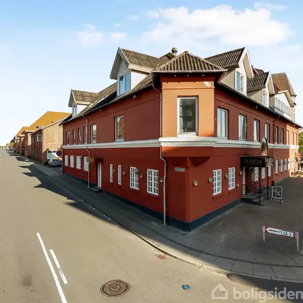 En stor, rød bygning med hvide vinduer står på et gadehjørne. På fortovet er et skilt med ordet "BRAUN" ved indgangen. Bygningen er omgivet af en vej og nabo-bygninger.