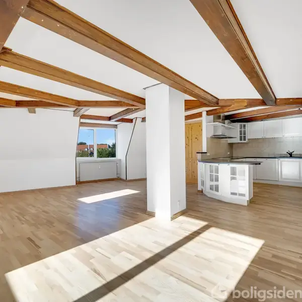 Et lyst og rummeligt loftsrum med træbjælker i loftet og parketgulv. Et åbent køkken med en hvælvet bardisk i forgrunden. Store vinduer bidrager med naturligt lys.