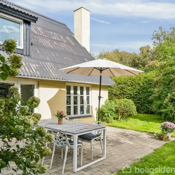 Et hvidt havebord med stole står under en parasol på en terrasse. Det er foran et gult hus omgivet af grønne buske og en velplejet græsplæne.