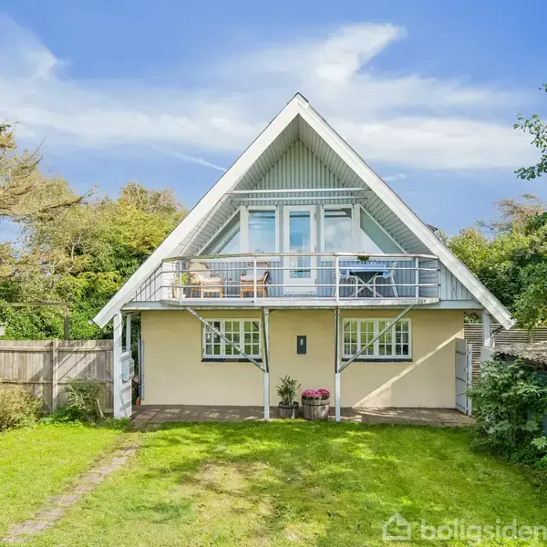 Et trekantet hus med altan står i en velplejet have. Huset har træbeklædning og store vinduer, omkranset af træer og en blå himmel i baggrunden.