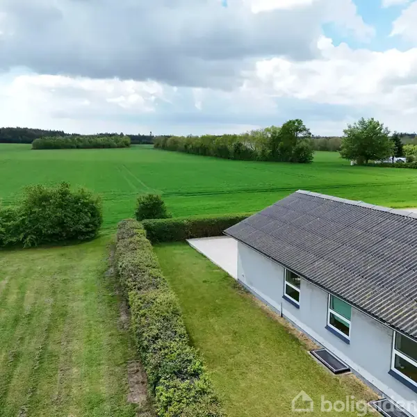En lys bygning med sort tag står langs en græsplæne med hække. Baggrunden viser en vidstrakt grøn mark og skov i horisonten under en skyet himmel.