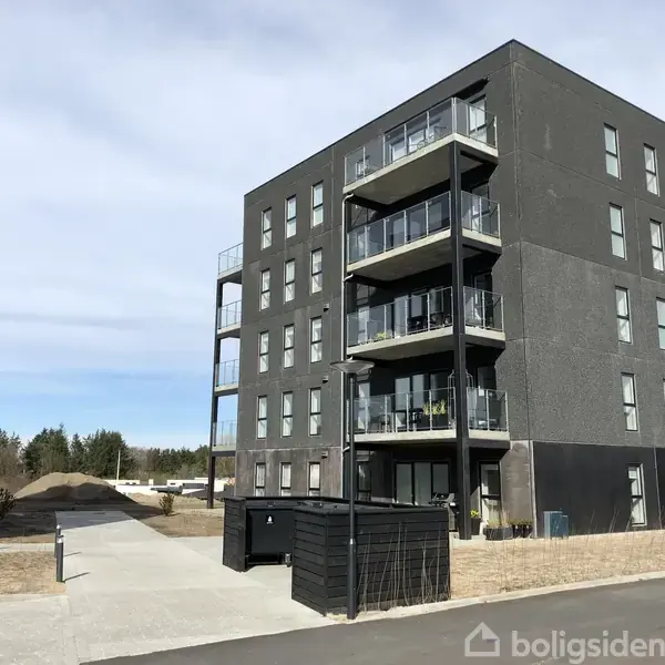 En fem-etagers moderne lejlighedsbygning med balkoner troner over en asfalteret sti i et boligområde med træer og åbne arealer.