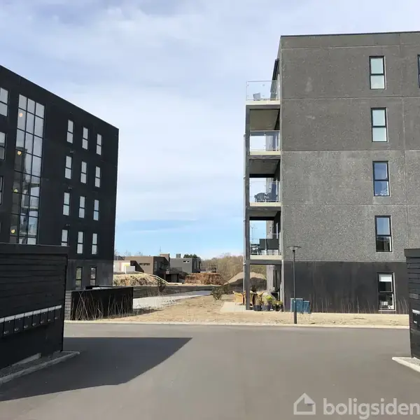 To sorte lejlighedsbygninger står overfor hinanden med flere vinduer. Mellem bygningerne er der en indgang til et gårdområde med træer. Fortove løber langs begge bygninger.