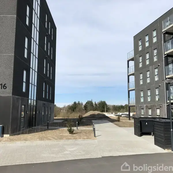 Sort lejlighedsbygning med tallet 16 på facaden står i stille boligkvarter ved siden af lignende bygning med altaner. Mellem bygningerne ses en sti og grønne områder.