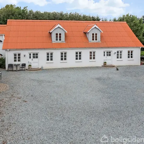 Et hvidt hus med rødt tegltag står på en gårdsplads dækket af grus, omgivet af frodig vegetation. Terrasse med bord og stole placeret ved siden af hoveddøren.