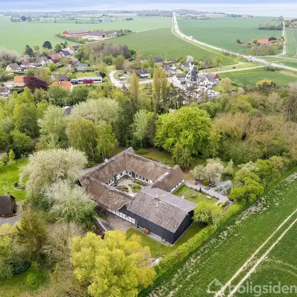 Et firkantet gårdkompleks med stråtage omgivet af frodig skov og marker. Det ligger i en landlig landsby med spredte huse og veje, der strækker sig gennem det grønne landskab.