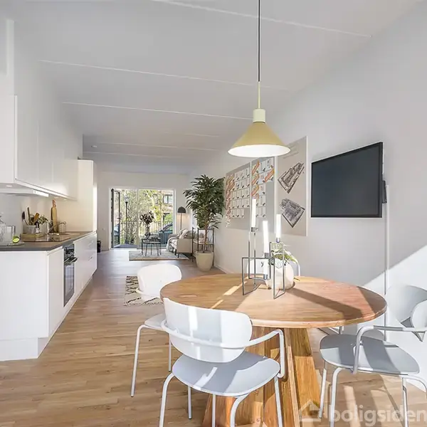 Rundt spisebord med fire stole står i en moderne, lys stue-køkken kombination. Et TV hænger på væggen, og glasskydedøre fører ud til en grøn have.