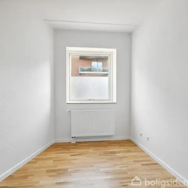 Et tomt rum med hvidmalede vægge og lyst trægulv. Et vindue med en radiator nedenunder udsigt til en bygning udenfor. Ingen tekst er synlig.