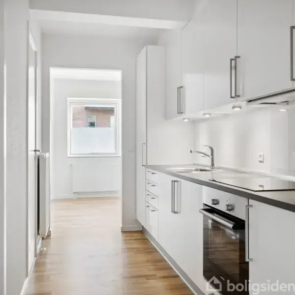 Et moderne køkken med hvide skabe og ståludstyr strækker sig langs væggen, mens et vindue i baggrunden giver naturligt lys ind i det lyse rum med trægulv.