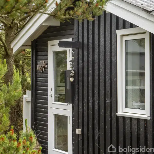 Et sort, træbeklædt sommerhus står stille blandt tætstående grantræer. Huset har hvide karme omkring et vindue og en glasdør, der åbner ind til en hyggelig atmosfære.