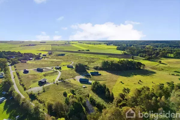 Landområde med spredte huse, der står roligt blandt marker og skove. Sollys skinner over det åbne, grønne landskab, og en snoet vej forbinder de forskellige bygninger.