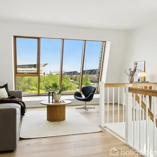 Køkken med hvidt interiør og trægulv, set fra siden af en åben skuffe. Spisebord med stole står foran foldedøre, der åbner mod en terrasse med udsigt over grønne områder.